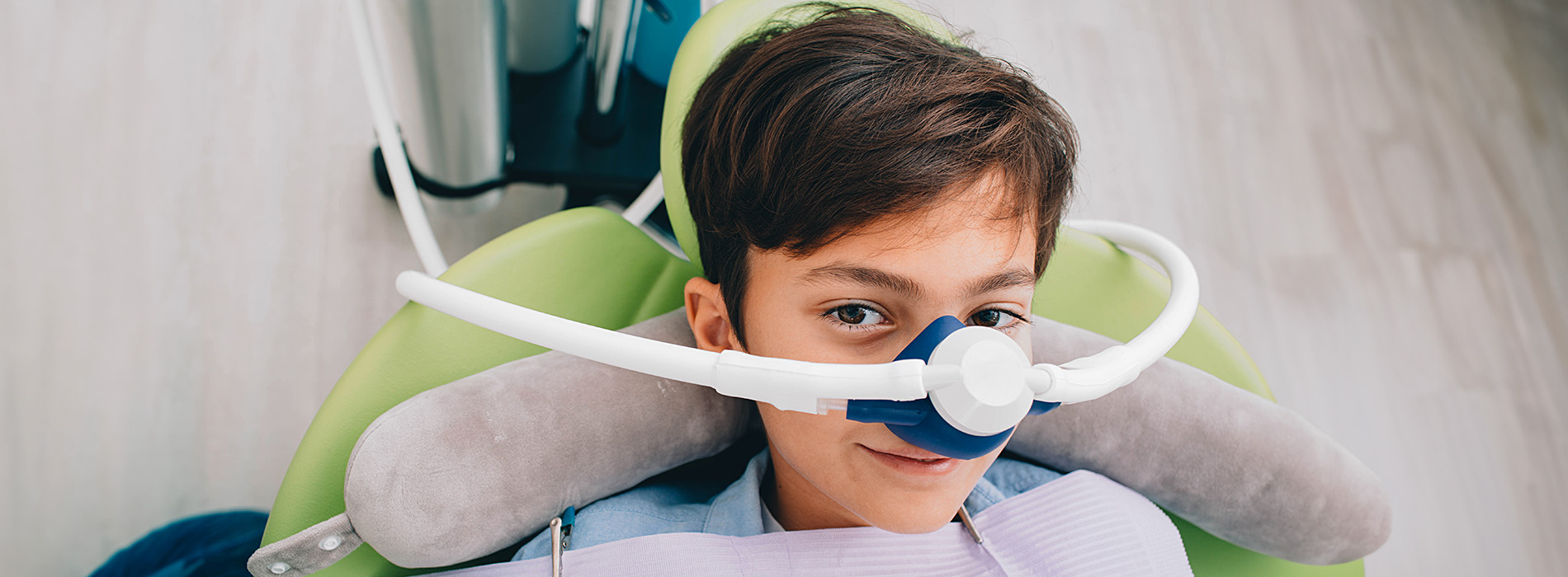 A young boy wearing a medical oxygen mask sits in a dental chair with a blue background.