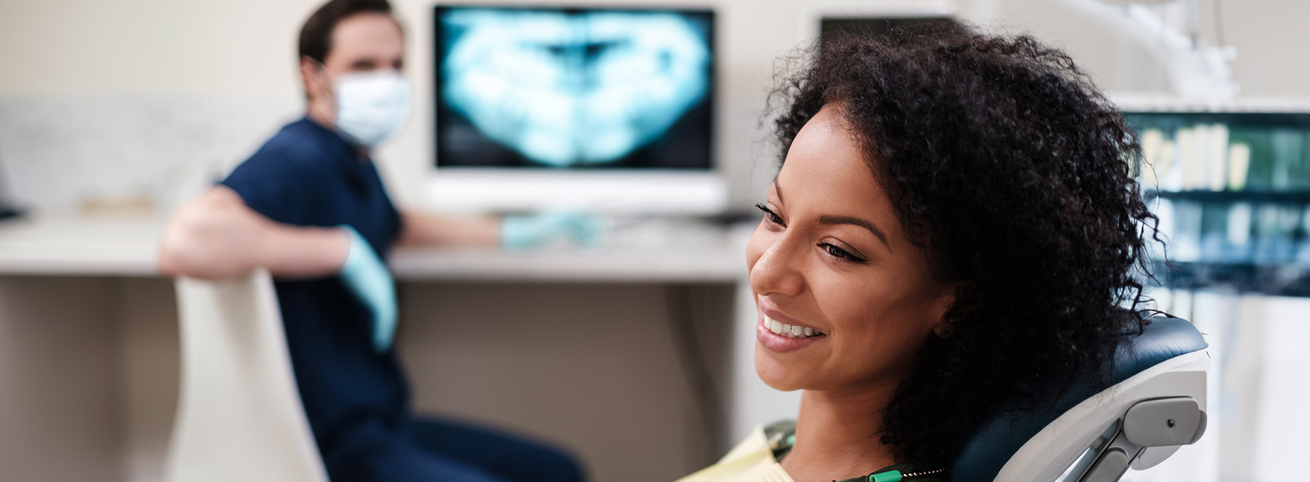 A person is seated in a dental chair, receiving care from a dental professional who stands behind them.