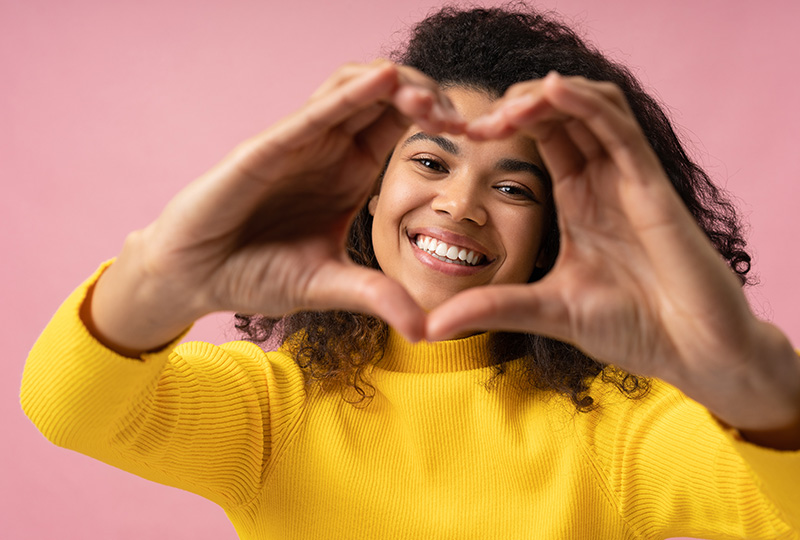 A woman with dark hair is making a heart shape with her hands while smiling at the camera.