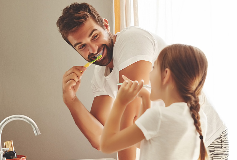 A man brushing his teeth while a child watches, both in front of a bathroom sink.