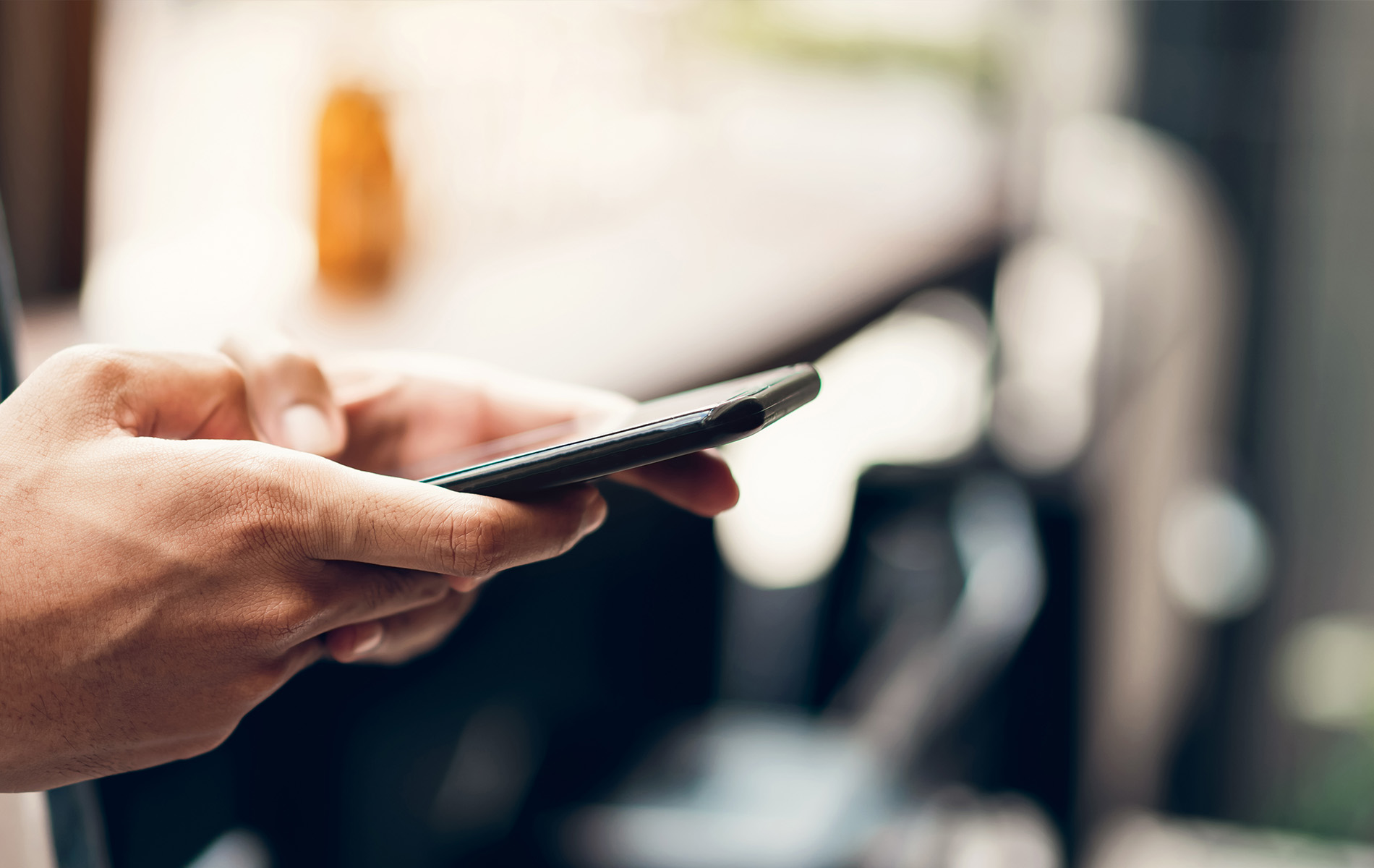 The image shows a person holding a smartphone with both hands, seemingly engaged in using it while standing in front of a blurred background that includes an urban setting with tables and chairs, suggesting a cafe or restaurant environment.