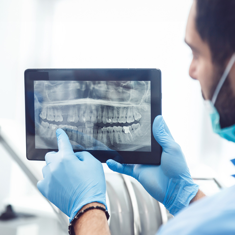 The image shows a dental professional using a tablet to view an X-ray of a patient s teeth while wearing protective gloves and a face mask, indicating a focus on hygiene and safety during the COVID-19 pandemic.