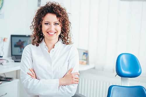 A woman with curly hair is standing in an office setting, smiling at the camera, wearing a white shirt and dark pants, positioned between two dental chairs.