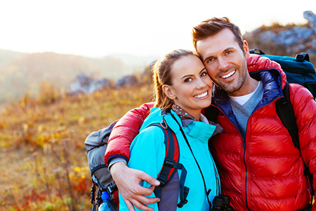 The image depicts a man and woman posing together outdoors during daylight hours, with both individuals wearing backpacks and smiling at the camera.