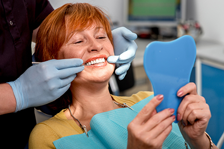 A woman wearing a blue surgical mask and holding a toothbrush-shaped object with fake teeth over her mouth, presumably for dental hygiene demonstration, with a smiling dentist in the background.