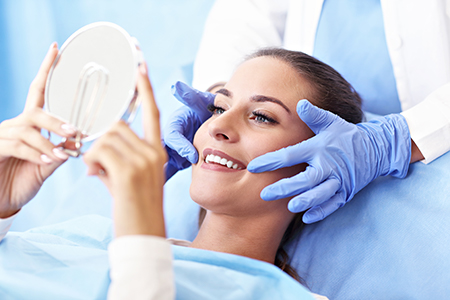 A woman seated on a dental chair with a mirror held up to her face, smiling at the camera, while a dentist or hygienist is taking a picture of her smile with a smartphone.