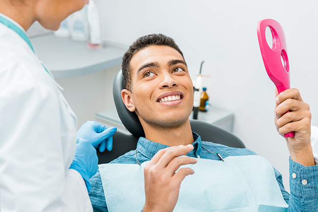 A man with a wide smile sitting in a dental chair while holding a pink toothbrush, receiving care from a dental professional.