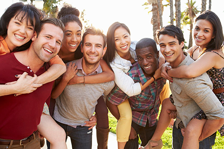 The image shows a group of people posing together outdoors during daylight, with one person holding up another for a hug, all smiling and appearing happy.