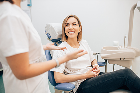 The image shows a woman sitting in a dental chair with a smile on her face, being attended to by a dental professional who appears to be performing a cleaning or examination.