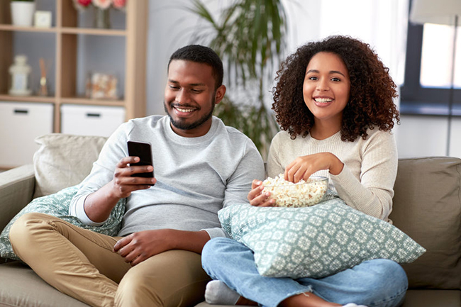 A man and woman sitting on a couch, smiling and enjoying popcorn while watching something together on a television screen.